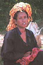 to 27 K Jpeg 9809H15 Pa'O woman sitting by her produce in Kalaw Market, Shan State. Note her turban tied from a woven scarf bought in the market and her Shan bag in her lap. She is wearing a short jacket, with long sleeves over her shift blouse which has a trim of embroidery in the 'V' of the neck and it is possible to see an echoing embroidery in the side seam of her blouse to the left of the photograph. The jacket has machine stitched detail around the piecing.