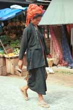 to 26K Jpeg 9809K01 Pa'O woman striding through the 5-day rotating market in Kalaw, Shan State. Her garments of black serge short, long-sleeved jacket with selvedge woven trim inset down the opening of the jacket, loose blouse with embroidered trim along the seams and calf length longyi are set off by the plaid scarf wound around her head and her striped bag