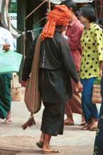 to 28K Jpeg 9809K02 Pa'O woman striding through the 5-day rotating market in Kalaw, Shan State. Her garments of black serge short, long-sleeved jacket with selvedge woven trim inset down the centre-back of the jacket, loose blouse with embroidered trim along the seams and calf length longyi are set off by the plaid scarf wound around her head and her striped bag 