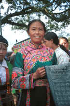 Jpeg 78K Batik artist shows her beautifully executed batik skirt length which indicates by its two intensities of indigo dye that it has been waxed twice.  I bought this length for my collection with the help of the school girl on the left who helped us barter by writing and interpreting the figures for the older lady.  Note the embroidered sleeves which can be removed and are placed over the sleeve of the jacket.  Da Shu Jia village, Xin Zhou township, Longlin county, Guangxi province 0010i18.jpg