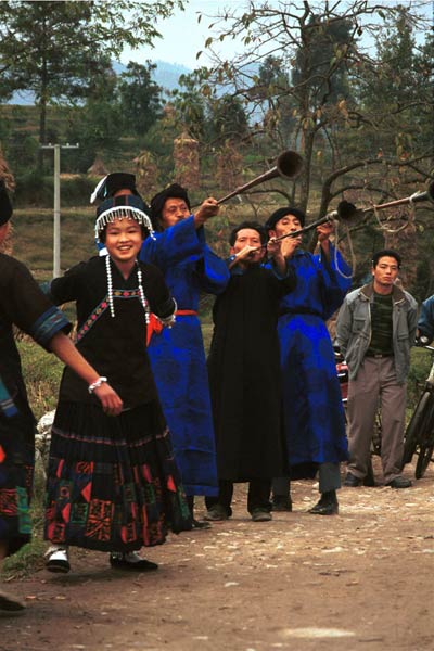 Jpeg 52K  0110D22  Girls dancing and trumpeters playing as part of our welcome to Gan He village, Ya Rong township, Huishui county, Guizhou province, south-west China. The people living in this village are known as Qing Miao. There has been much intermarriage between Miao and Bouyei which is evident from the every-day costume of the women. The pieced skirts of applique and wax-resist being worn by the girls were originally only worn at funerals but today they are worn for special festivals and dance competitions.