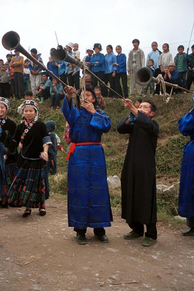 Jpeg 52K  0110D28 Girls dancing and very long and loud trumpets being played as part of our welcome to Gan He village, Ya Rong township, Huishui county, Guizhou province, south-west China. The people living in this village are known as Qing Miao. There has been much intermarriage between Miao and Bouyei which is evident from the every-day costume of the women. The pieced skirts of applique and wax-resist being worn by the girls were originally only worn at funerals but today they are worn for special festivals and dance competitions.