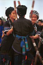 to Jpeg 57K  0110D36 Girls beating sticky rice as part of a dance programme in Gan He village, Ya Rong township, Huishui county, Guizhou province, south-west China. The people living in this village are known as Qing Miao. There has been much intermarriage between Miao and Bouyei which is evident from the every-day costume of the women. The pieced skirts of applique and wax-resist being worn by the girls were originally only worn at funerals but today they are worn for special festivals and dance competitions. Note the bands of complex weaving on the sleeves of the women's jackets.