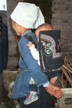 to Jpeg 56K  0110E16 A baby held in an attractive baby carrier with embroidery and applied braid in Gan He village, Ya Rong township, Huishui county, Guizhou province. This is a Qing Miao (or Bouyei/Miao as there has been much intermarriage and the costume is very mixed). 