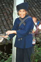 to Jpeg 50K 0110E17  An old woman with a baby in a baby carrier on her back holding the ends of warp threads which are being untangled and straightened before warping up a loom in Gan He village, Ya Rong township, Huishui county, Guizhou province. This is a Qing Miao (or Bouyei/Miao as there has been much intermarriage and the costume is very mixed).