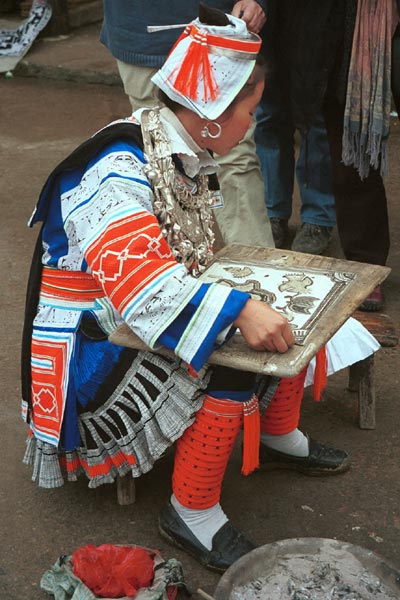 Jpeg 60K 0100F32  Gejia woman dressed in full festival finery demonstrating wax resist. The design on her own costume - her headdress and jacket at least (the black on white) - is probably all made from a commercially printed fabric and is not made from traditional wax resist. Ma Tang village, Kaili City, Guizhou province.