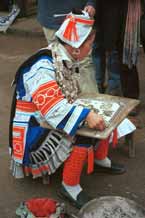 to Jpeg 60K 0100F32  Gejia woman dressed in full festival finery demonstrating wax resist. The design on her own costume - her headdress and jacket at least (the black on white) - is probably all made from a commercially printed fabric and is not made from traditional wax resist. Ma Tang village, Kaili City, Guizhou province.