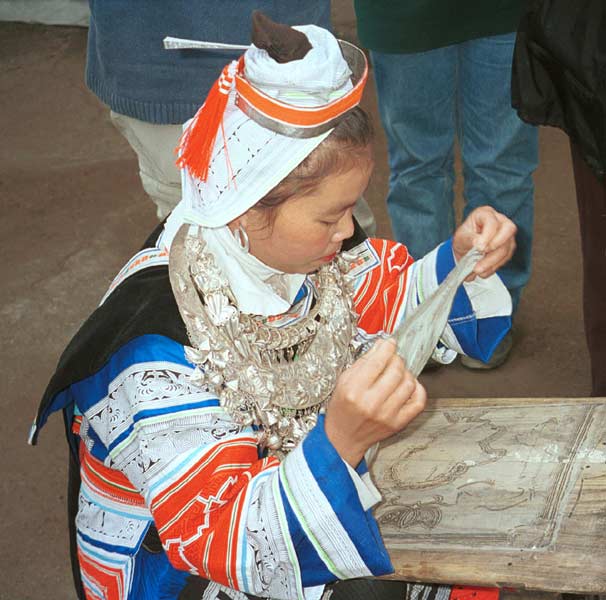 Jpeg 56K 0100F33 Gejia woman dressed in full festival finery about to fix a piece of cotton fabric to a board before starting to wax a design. The design on her own costume - the black on white - is probably all made from a commercially printed fabric and is not made from traditional wax resist. Ma Tang village, Kaili City, Guizhou province.