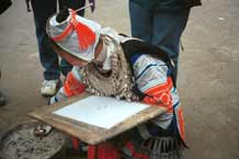 to Jpeg 58K 0100F34 Gejia woman dressed in full festival finery demonstrating wax resist. The design on her own costume - her headdress and jacket at least (the black on white) - is probably all made from a commercially printed fabric and is not made from traditional wax resist. She has heated the wax in a metal dish on hot ashes. Ma Tang village, Kaili City, Guizhou province.