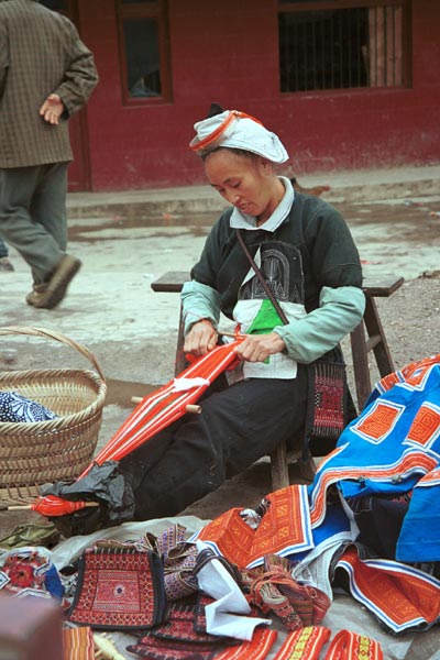 Jpeg 58K 0111G25  Gejia woman weaving a braid by a heap of embroidered clothing, woven braids and also some bags from a local Miao (not Gejia) group. Ma Tang village, Kaili City, Guizhou province.