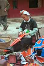 to Jpeg 58K 0111G25  Gejia woman weaving a braid by a heap of embroidered clothing, woven braids and also some bags from a local Miao (not Gejia) group. Ma Tang village, Kaili City, Guizhou province.