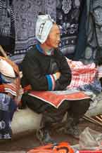 to Jpeg 57K 0111G30 Gejia woman sitting amongs her modern wax resist work made to sell to the tourists. Note her embroidered apron which is edged with some original wax resist. Her headdress is, however, made from a commercially manufactred fabric using traditional Gejia designs. Ma Tang village, Kaili City, Guizhou