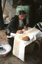 Jpeg 29K Young Side comb Miao woman sitting waxing the batik which will be indigo dyed to go at the top of a skirt.  She is copying the batik from another skirt and has a lump of bees wax in front of her on which she wipes excess wax off her waxing instrument and a pot of melted wax sitting in a bowl of hot embers beside her.  Long Dong village, De Wo township, Longlin country, Guangxi province 0010e21.jpg