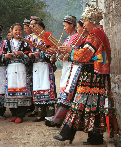Jpeg 123K Miao women playing lusheng pipes to welcome us. The two girls to the left are wearing the modern festival costume of the village and are holding carved charms on threads to put over out heads. The woman near to the camera on the right has a skirt and silver head dress which is not local to this area. It may have been adopted to create more of a stir in the dance competitions or she might have maried into the village from elsewhere. Lou Jia Zhuang village, Anshun city, Guizhou province. 0110A20