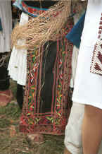 Jpeg 35K Front view of a White Miao woman showing her machine embroidered apron over a pleated hemp or ramie woven skirt over which is draped hemp or ramie fibre stripped from the bush which she is splicing as she stands and waits - Ma Wo village, Zhe Lang township, Longlin county, Guangxi province 0010j37.jpg