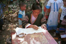 Jpeg 44K Side Comb Miao married woman working on a batik strip for the top of a skirt.  See the on the ground to the left of the photo her wax warming in hot embers and the slab of wax on the table on which she wipes the excess off her batik instrument.  Pao Ma Cheng village, Teng Jiao township, Xingren country, Guizhou province. 0010n21.jpg