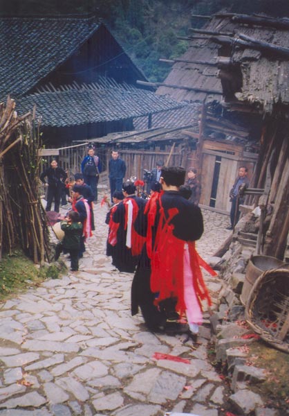 Jpeg 57K e30e 25 February 2004, the fourth day of the festival, in Langde village, Guizhou province, On this day, the ceremony was to take the spirit of the mountain dragon inside each home to live there so the family would enjoy good fortune in the future. The shaman and a group of men all wore traditional dress. These men were the leading people of the ceremony and of the community. They must be well thought of by the villagers before they can be selected as participants or else they cannot play the roles. When the ceremony started, the shaman went in front leading the way to each home. On entering each family's house they were offered cups of rice wine by the head of the family and fire crackers were also set off. While in the house, they played lusheng and danced in a circle to the rhythm of the bronze drum. When leaving the house, the owner gave them some red ribbons and cigarettes. 