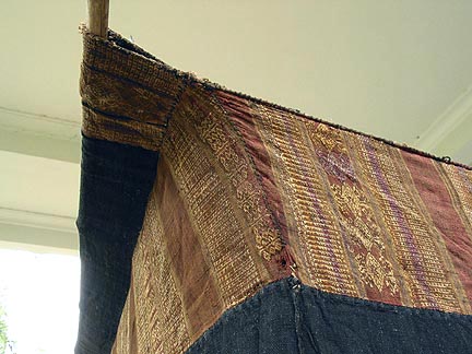 Jpeg 52K Top corrner hanging detail of Tai Daeng Mosquito Net (pha mung) from Xam Tai in northeast Laos. Approximately: 61" x 65" on the sides and 60" high; hanging pockets extends 4" from main 'box'. Four panels on each side of handspun, handwoven indigo cotton, with silk and cotton heading border in good condition; top of four panels (one is pieced making three parts) gold-colored handspun, handwoven cotton. One side (side 'd') has a panel replaced, or of darker color indigo. Several patches and some holes, although none are larger than 1". A few patches of supplementary warp scraps. All handstitched. Early 20th century.