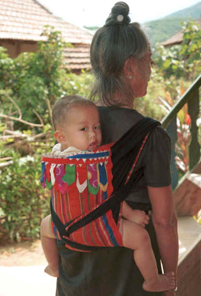 Jpeg 27K Black Thai baby being carried by its grandmother's back.  The baby carrier is made with an insertion of hand woven supplementary weft fabric (probably cotton ground and silk supplementary wefts) and adorned with strips of inserted folded 'points' and hanging tabs of different bought fabric scraps which have been carefully sewn together and turned.  Tuan Giao on the road from Son La to Dien Bien Phu in Son La Province.  9510C32.JPG