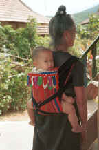to Jpeg 27K Black Thai baby being carried by its grandmother's back.  The baby carrier is made with an insertion of hand woven supplementary weft fabric (probably cotton ground and silk supplementary wefts) and adorned with strips of inserted folded 'points' and hanging tabs of different bought fabric scraps which have been carefully sewn together and turned.  Tuan Giao on the road from Son La to Dien Bien Phu in Son La Province.  9510C32.JPG