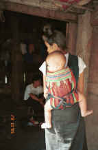 to Jpeg 26K Black Thai baby being carried on it's grandmother's back in a baby carrier made with an insertion of supplementary woven cloth and layers of edging made from individually folded 'points'.  A village near Dien Bien Phu, Lai Chau Province. 9510E21.JPG