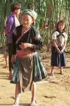 to Jpeg 44 K  9510G2Older Flowery Hmong woman with children outside a house in small road-side village near Phong Tho (Phong Thanh) in Lai Chau Province. Note her short pleated skirt with batik and apliqued fabric squares and also her long, indigo apron.  Her head cloth will have been bought in the market - machine woven and imported from China.