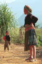 to Jpeg 49K  9510G24 Older Flowery Hmong woman and child outside a house in small road-side village near Phong Tho (Phong Thanh) in Lai Chau Province. Note her short pleated skirt with batik and apliqued fabric squares and also her long, indigo apron.  Her head cloth will have been bought in the market - machine woven and imported from China