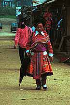 to Jpeg 48K 9510K26  Brightly dressed Flowery Hmong woman walking down the street in Sa Pa, Lao Cai Province.  She is a visitor to Sa Pa, in the town on a training programme, and lives near the Chinese border. The Flowery Hmong near to the border tend to wear more bought trimmings on their clothing which they obtain from China.  Note her embroidered and applique leg wrappings and her long apron.  She only very narrow bands of wax resist on her skirt which features applied strips of fabric and embroidery. 