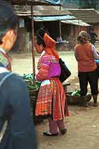 Jpeg 42K 9510K28  Brightly dressed Flowery Hmong woman at a shop stall in Sa Pa, Lao Cai Province.  She is a visitor to Sa Pa, in the town on a training programme, and lives near the Chinese border. The Flowery Hmong near to the border tend to wear more bought trimmings on their clothing which they obtain from China.  Note her embroidered and applique leg wrappings and her long apron.  She only very narrow bands of wax resist on her skirt which features applied strips of fabric and embroidery. 