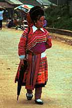 to Jpeg 48K 9510K29  Brightly dressed Flowery Hmong woman walking down the street in Sa Pa, Lao Cai Province.  She is a visitor to Sa Pa, in the town on a training programme, and lives near the Chinese border. The Flowery Hmong near to the border tend to wear more bought trimmings on their clothing which they obtain from China.  Note her embroidered and applique leg wrappings and her long apron.  She only very narrow bands of wax resist on her skirt which features applied strips of fabric and embroidery. 