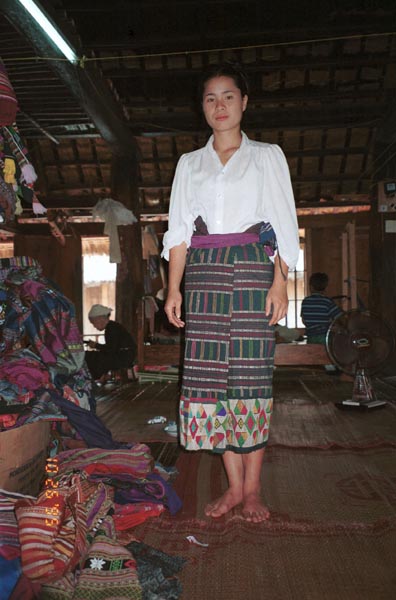 An old Thai minority woven sarong being modeled by a Thai girl in a village West of Hanoi, Vietnam.  The sarong is now in my collection.  See the old lady in the background winding bobbins ready for weaving.  In the foreground is a pile of woven lengths from various families in the village ready for sale. 9510A14E